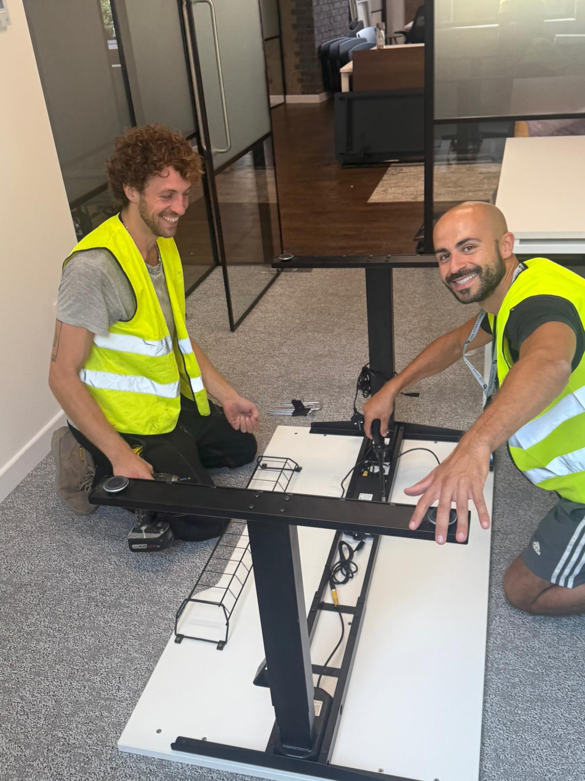 Two OfficeMatrix team members in hi-vis vests smiling while assembling a height-adjustable desk in a London office