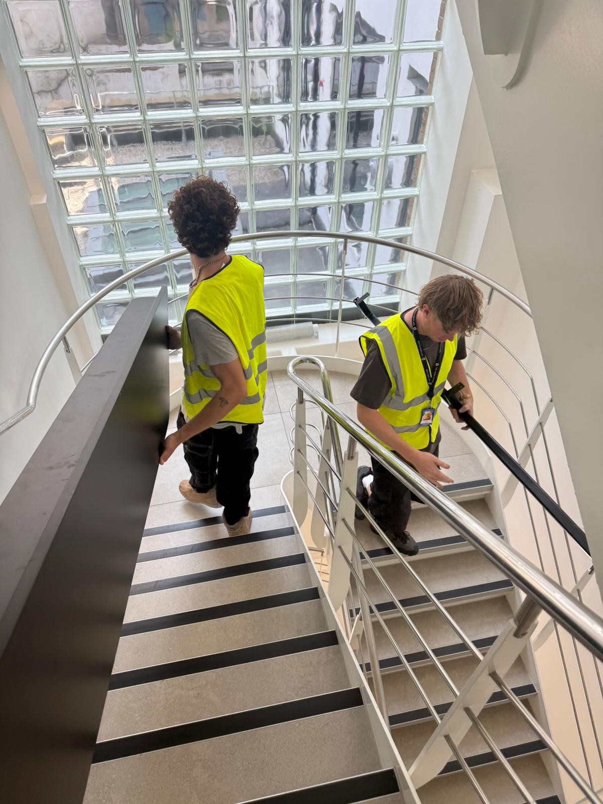 Two OfficeMatrix team members in hi-vis vests carrying a desktop panel down a bright spiral staircase beside a glass-block wall